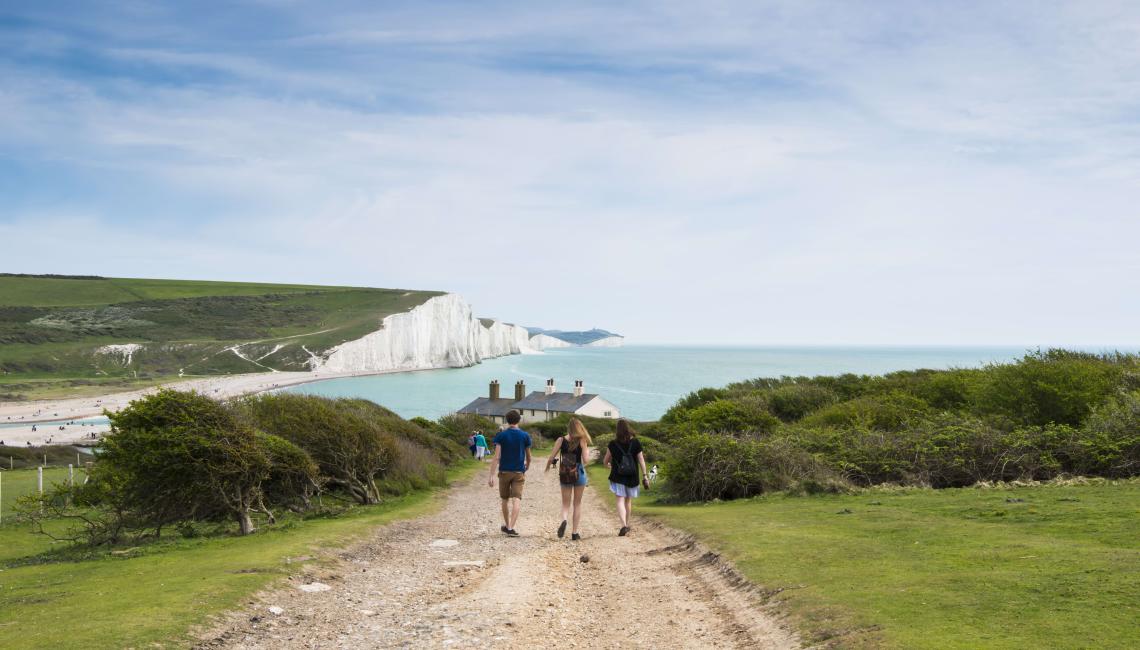 Walkers on a coastal path heading towards white chalk cliffs and the sea