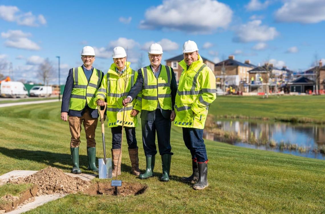 Four people in hard hats and hi-vis planting a young tree beside a pond and new homes