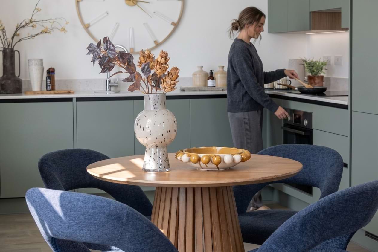 Woman cooking in a sage-green kitchen behind a round dining table with blue chairs